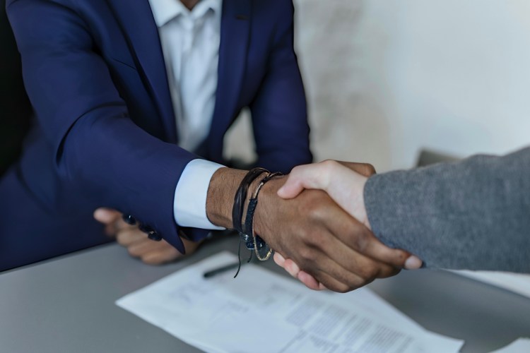 Photo of two people's arms with hands clasped in a handshake. In the background are papers on the desk.